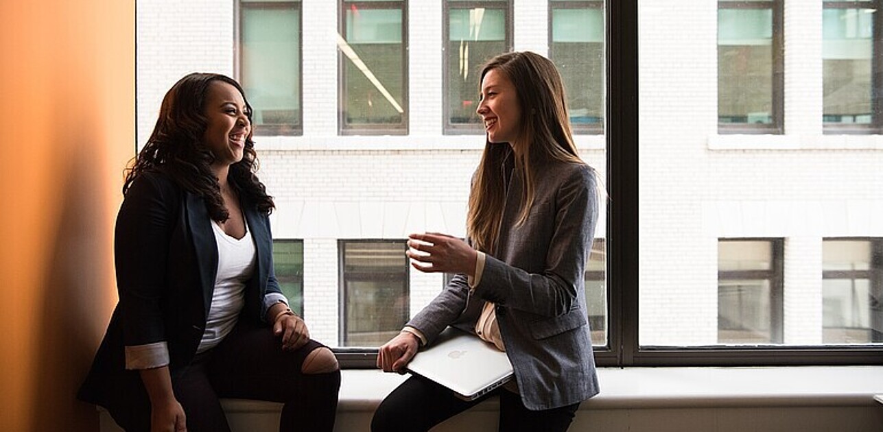 Two females dressed business casual sitting at a window talking to each other.