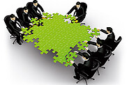 Elegant individuals seated around a table made of puzzle pieces, collaboratively adding more pieces to complete it.