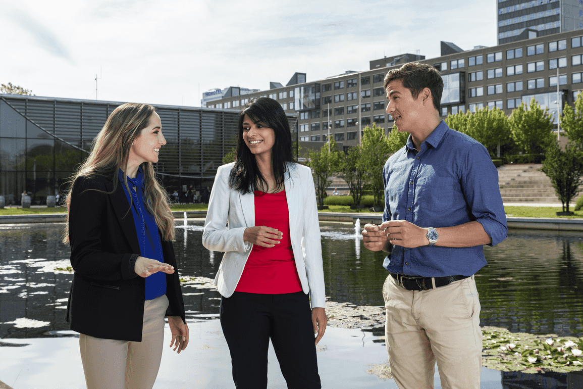 2 female and 1 male students are engaged in conversation with background of campus 