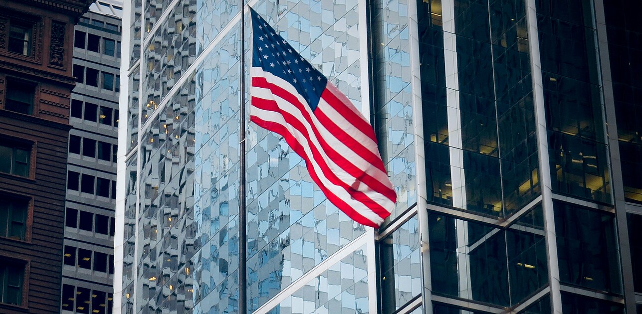 An American flag flying in front of glass office towers in a US business district, illustrating how US elections affect companies and stock markets.
