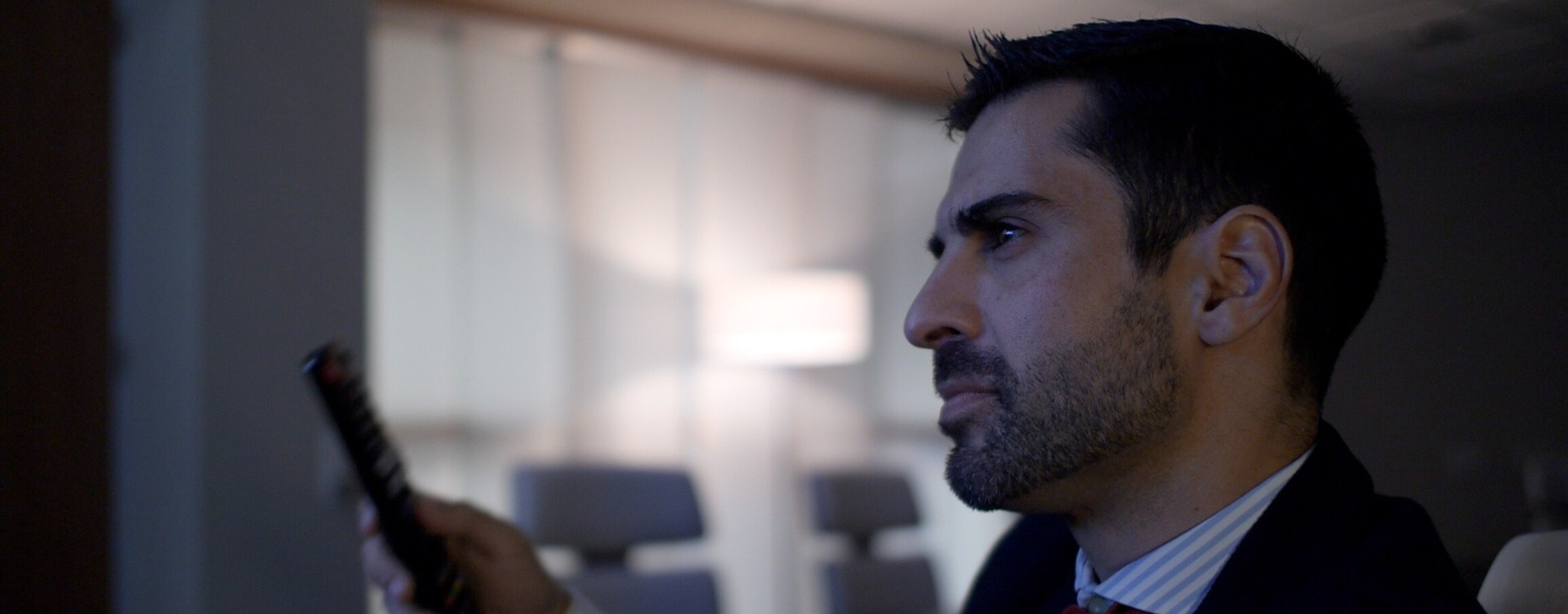 Close-up of a confident man in a suit, presumably a leader, alone in a conference room, holding a remote control, with a focused expression on his face.