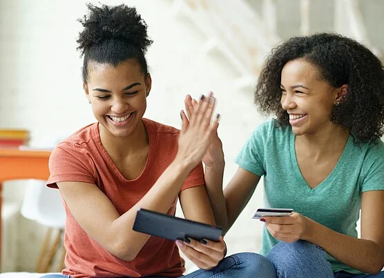 Two friends sitting together and smiling as they high-five, one holding a tablet and the other a bank card, illustrating how people share digital offers and rewards with each other.