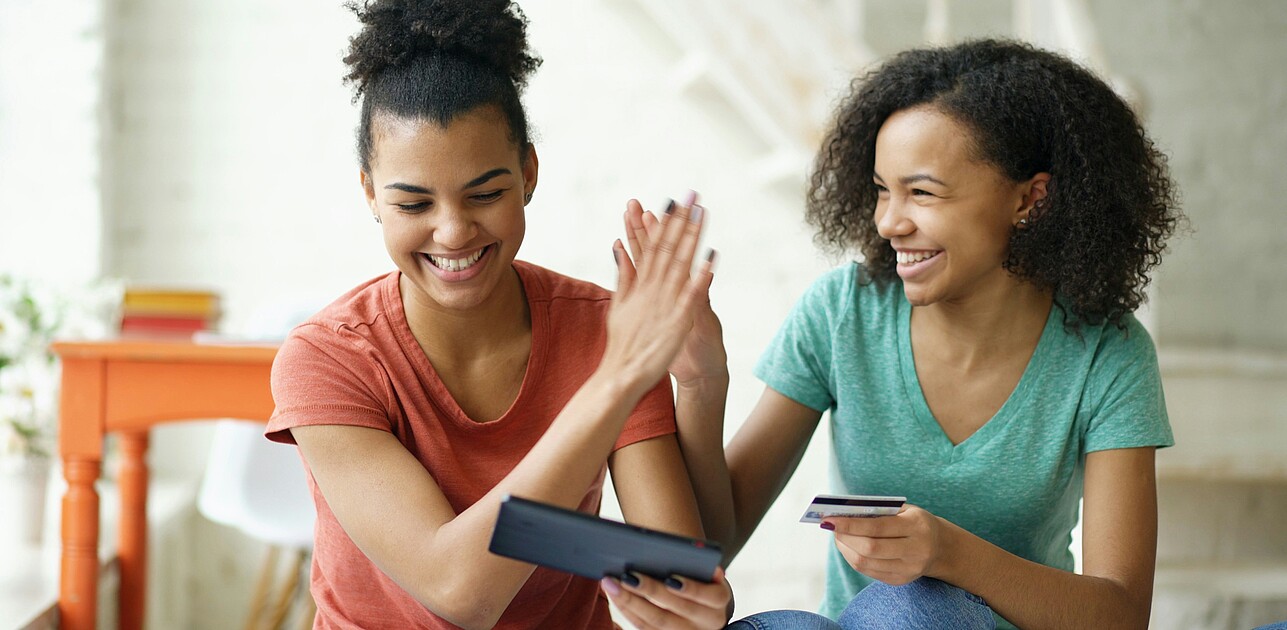 Two friends sitting together and smiling as they high-five, one holding a tablet and the other a bank card, illustrating how people share digital offers and rewards with each other.