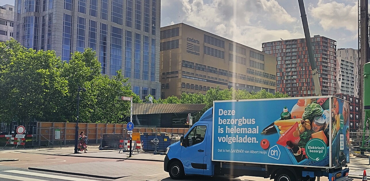 Delivery truck parked on a city street in Rotterdam with a large advertisement on its side. Tall office and residential buildings are visible in the background, along with trees and a pedestrian crosswalk in the foreground.