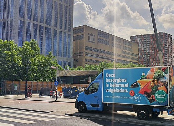 Delivery truck parked on a city street in Rotterdam with a large advertisement on its side. Tall office and residential buildings are visible in the background, along with trees and a pedestrian crosswalk in the foreground.