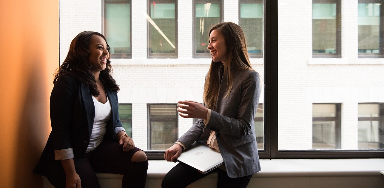 Two girls sitting on a windowsill, engaged in a cheerful conversation with smiles on their faces.