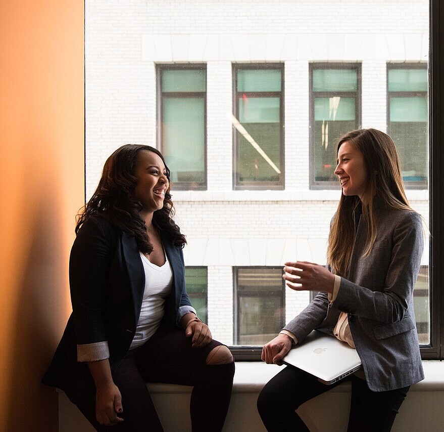 Two girls sitting on a windowsill, engaged in a cheerful conversation with smiles on their faces.