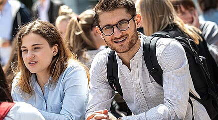 Two RSM students sitting outdoors among a group of other students, with one smiling at the camera and wearing glasses and a backpack.