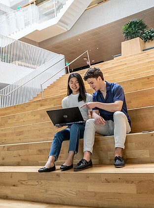 Two International Business Administration students, one holding a laptop and the other pointing towards the laptop, whilst sitting on the first floor staircase of the Polak building located on the Erasmus University campus