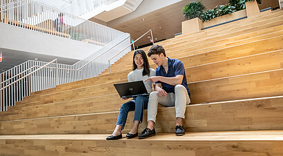 Two International Business Administration students, one holding a laptop and the other pointing towards the laptop, whilst sitting on the first floor staircase of the Polak building located on the Erasmus University campus