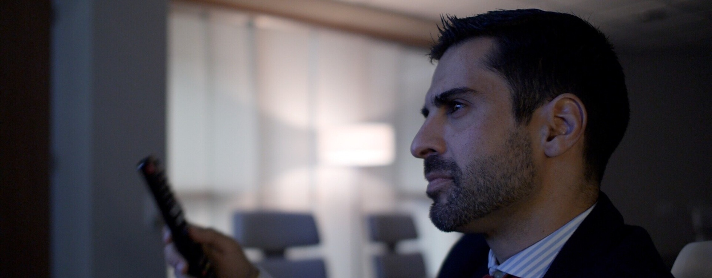 Close-up of a confident man in a suit, presumably a leader, alone in a conference room, holding a remote control, with a focused expression on his face.