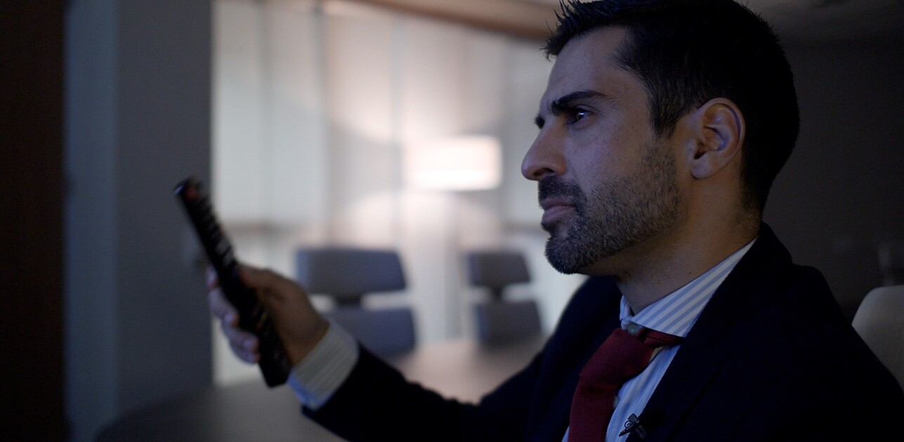 Close-up of a confident man in a suit, presumably a leader, alone in a conference room, holding a remote control, with a focused expression on his face.