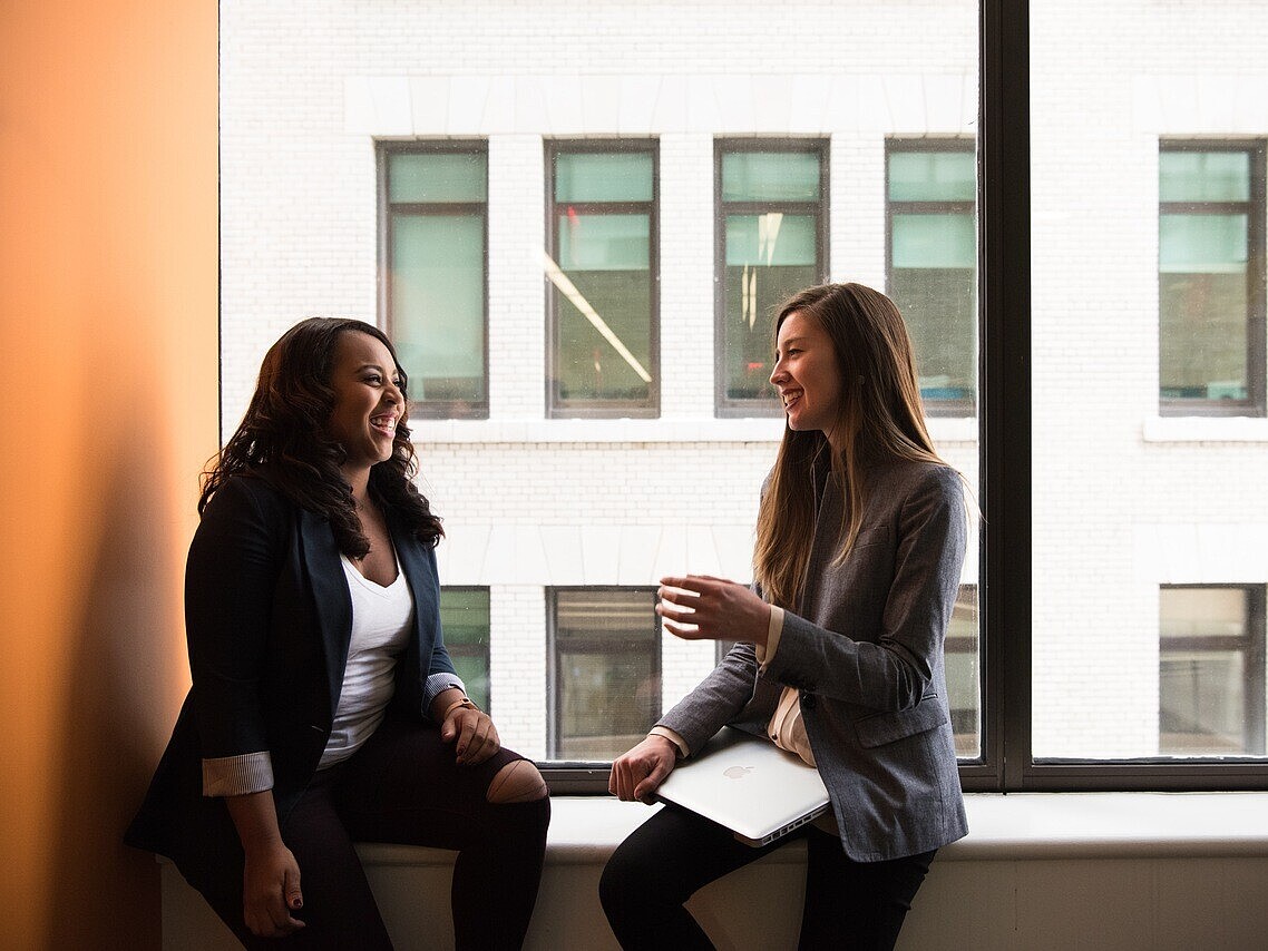 Two girls sitting on a windowsill, engaged in a cheerful conversation with smiles on their faces.