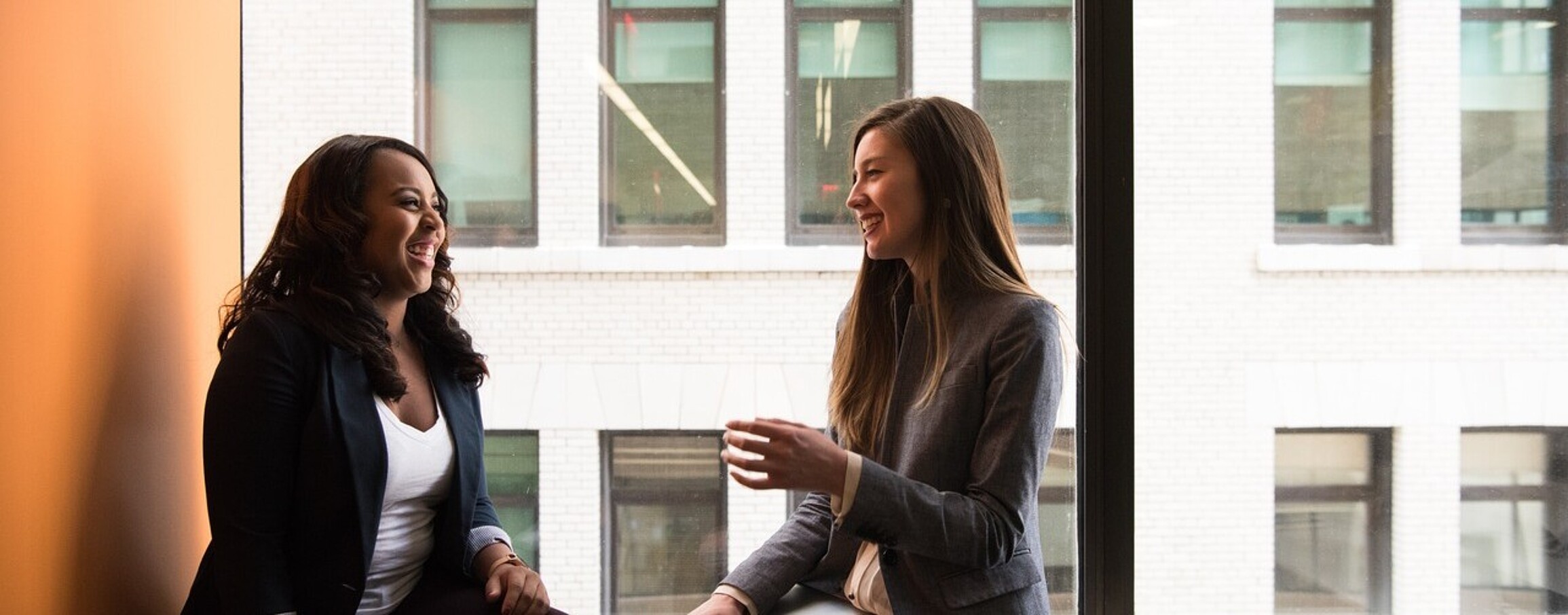 Two girls sitting on a windowsill, engaged in a cheerful conversation with smiles on their faces.