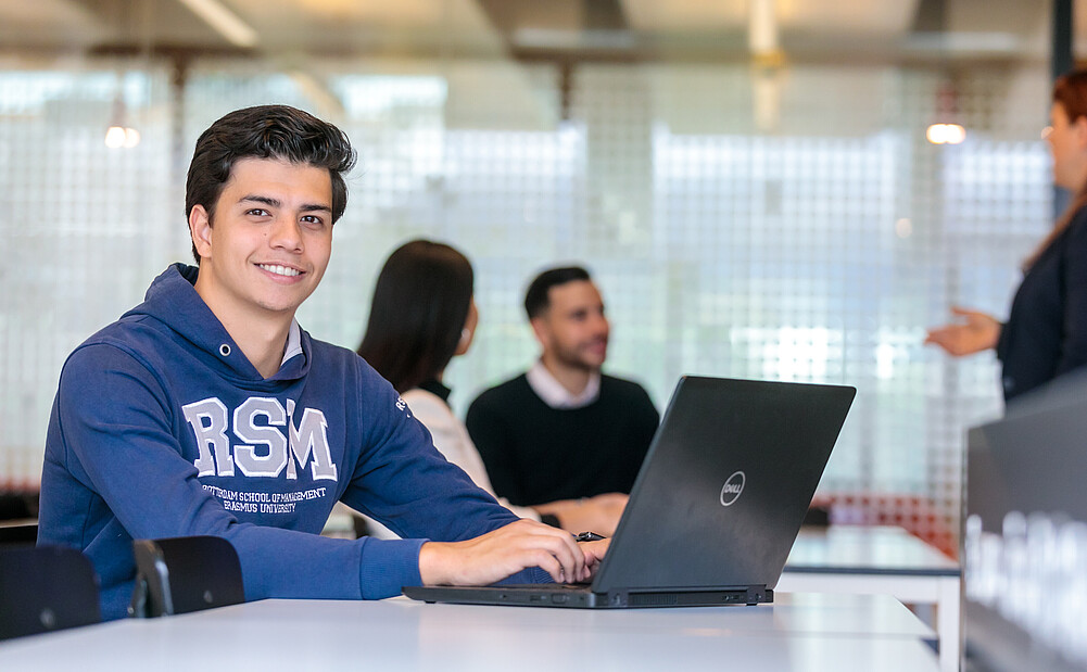 Enthusiastic Rotterdam School of Management student engaged in a workshop, wearing a cheerful smile, while diligently working on his laptop.