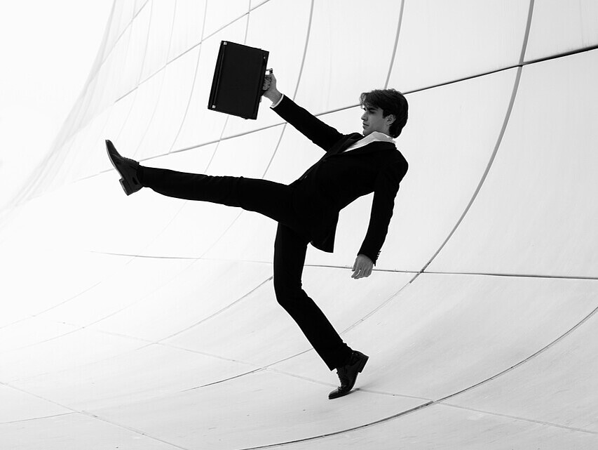 Young man in a suit and tie, holding a briefcase, making an exaggerated, comical step, juxtaposing professionalism with playful behavior.