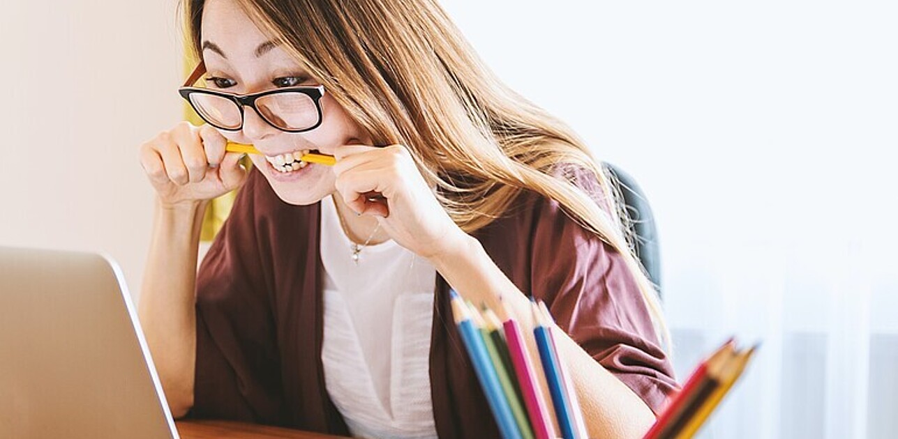 Woman wearing glasses bites on pencil whilst looking at computer scree.