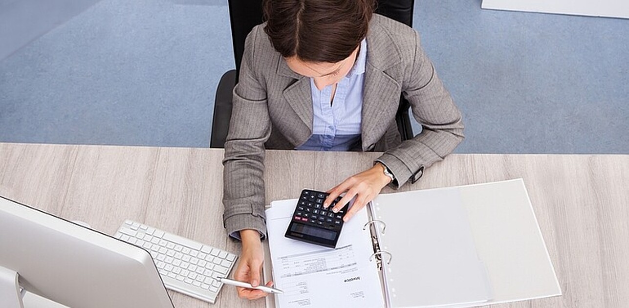 A lady is working at her office, reading through files and she is doing calculations with a calculator