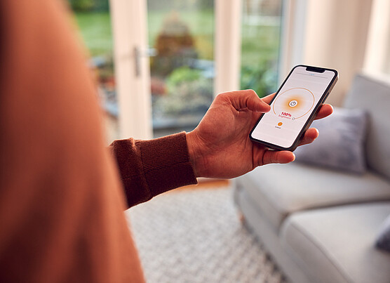 Person holding a smartphone with a home lighting control app displayed, adjusting brightness while standing in a modern living room.