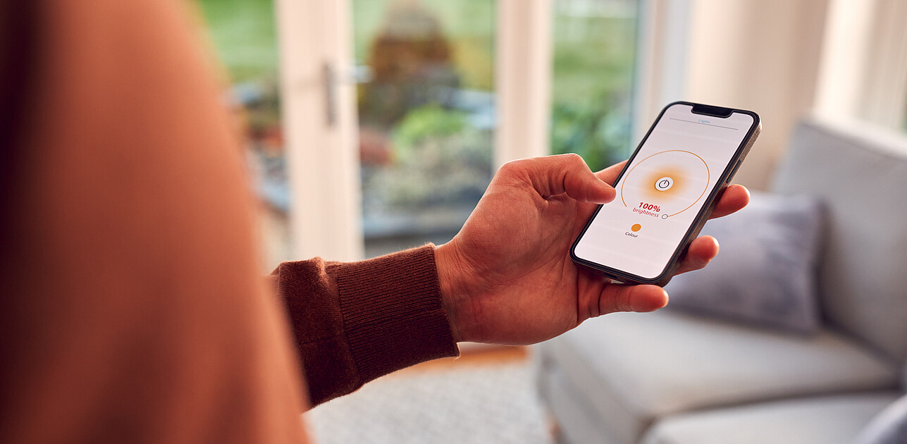 Person holding a smartphone with a home lighting control app displayed, adjusting brightness while standing in a modern living room.