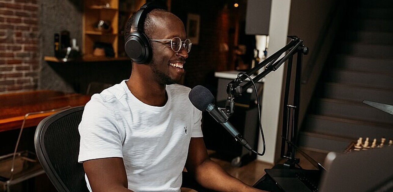 Male wearing headphones sitting in a recording studio talking into a microphone.
