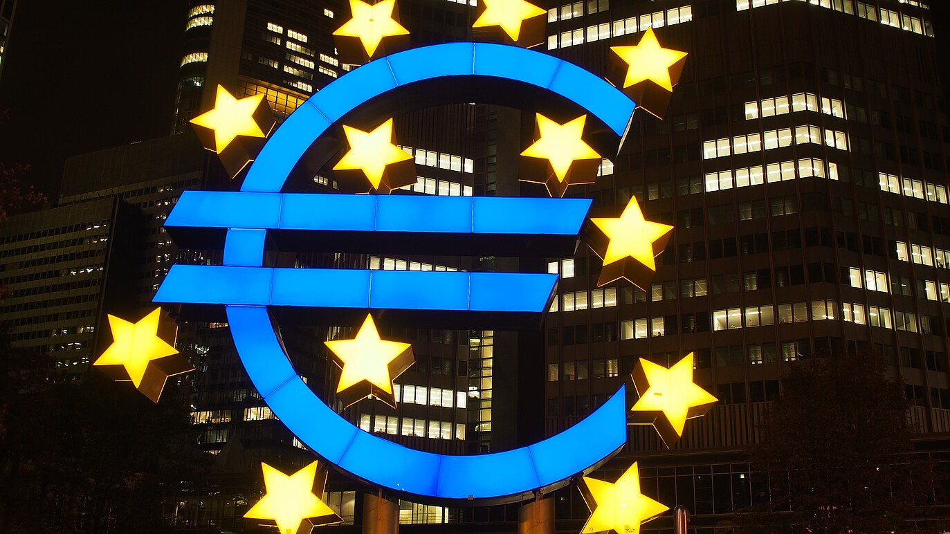 A photo shows illuminated euro currency sign with skyscrapers on the background 