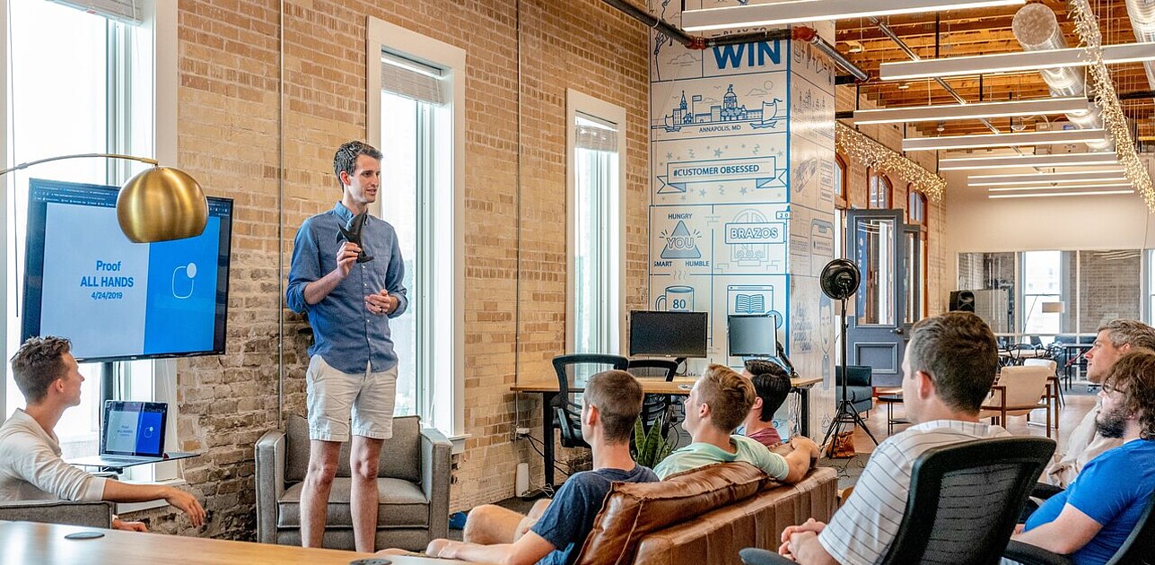 Young employee standing in front of a screen, giving a presentation to attentive colleagues in a meeting room.